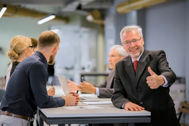 Bezpłatne zdjęcie uśmiechnięty starszy biznesmen pokazuje kciuk up podpisuje przed biznesmenami dyskutuje w biurze