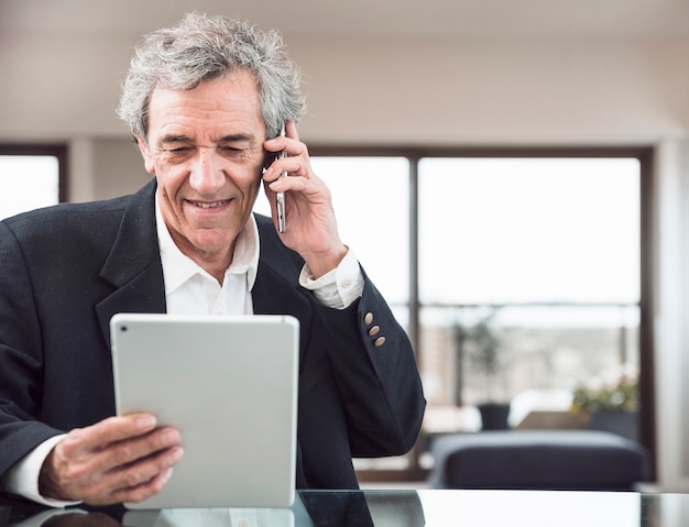 Uśmiechnięty starszy mężczyzna opowiada na telefonie komórkowym patrzeje cyfrową pastylkę przy miejscem pracy