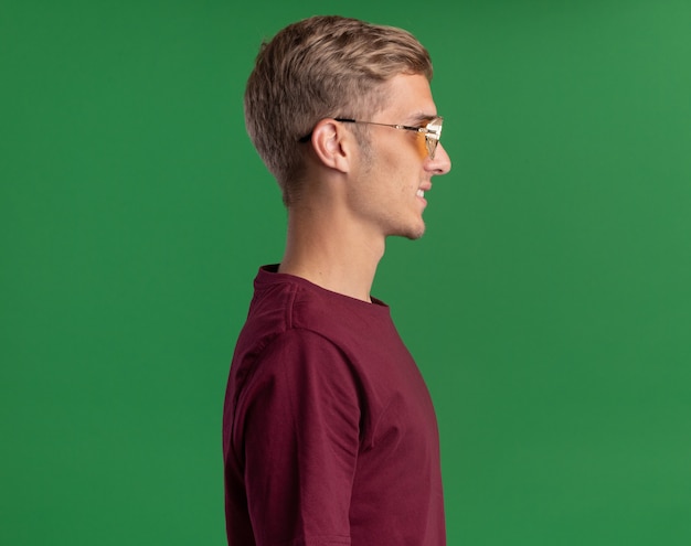 Uśmiechnięty stojący w widoku profilu młody przystojny facet w czerwonej koszuli i okularach odizolowanych na zielonej ścianie z kopią przestrzeni