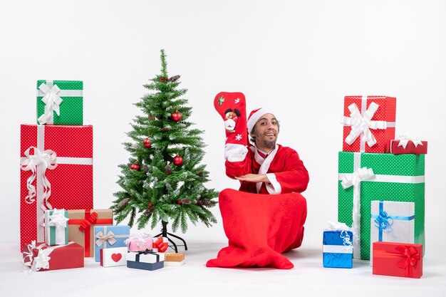 Uśmiechnięty święty mikołaj siedzi na ziemi i nosić skarpety świąteczne w pobliżu prezentów i udekorowane drzewo noworoczne na białym tle