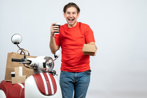 Uśmiechnięty szczęśliwy dostawy mężczyzna w czerwonym mundurze stojącym w pobliżu skutera pokazujący małe pudełko na białym tle