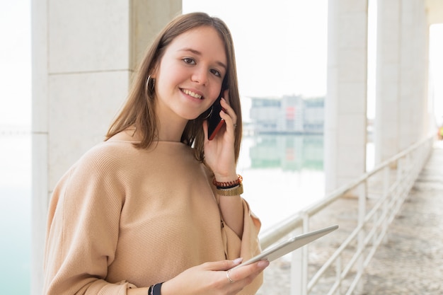 Uśmiechnięty żeński nastolatek używa telefon i pastylkę outdoors
