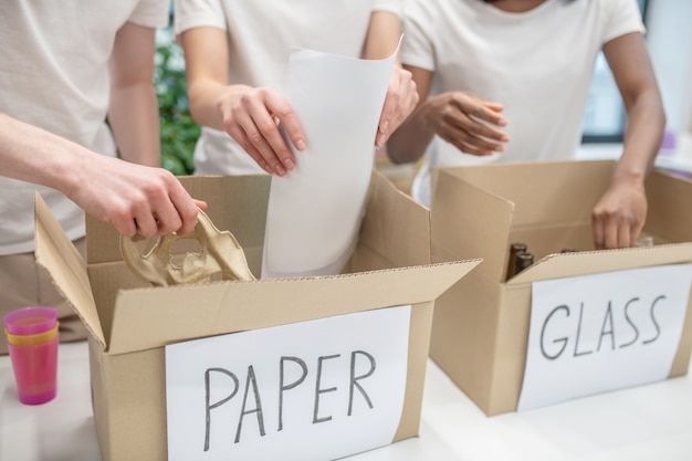 Utylizacja śmieci. ręce młodych ludzi w identycznych białych koszulkach sortują papier i szkło do pudełek do recyklingu