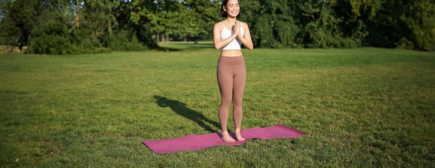 Bezpłatne zdjęcie uważność i dobre samopoczucie młoda kobieta uprawiająca jogę stojąca na macie w parku robiąca asany medytując na