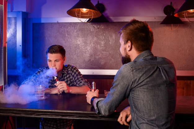 Vaping mężczyzna posiadający mod. Chmura pary na vapebarze. Dwóch mężczyzn odpoczywa w barze i pali elektroniczne papierosy.