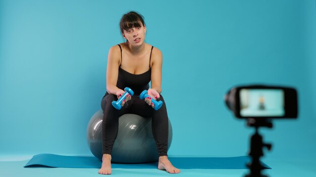 vloger fitness nagrywa wideo z treningu na kamerze w studio, używając hantli do wyjaśnienia techniki podnoszenia w praktyce sportowej. Muskularna kobieta filmowania lekcji szkolenia online na tonowanie piłki.