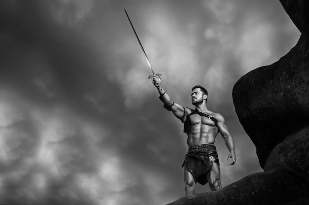 W imię Boga. Monochromatyczny portret potężnego, umięśnionego gladiatora trzymającego swój miecz pod chmurnym niebem copyspace