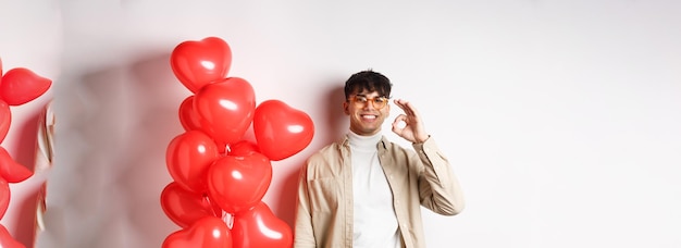 Bezpłatne zdjęcie walentynki i romans koncepcja szczęśliwy młody człowiek uśmiechający się pokazujący znak ok w pobliżu czerwonych serc gest recom