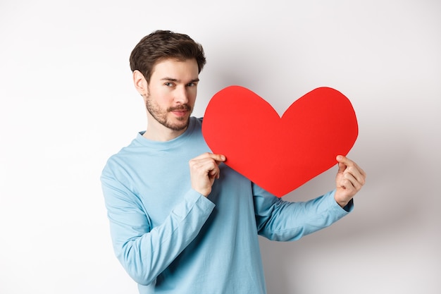 Walentynki. Przystojny i romantyczny mężczyzna trzyma duży czerwony valentine serce wyłącznik, patrząc uwodzicielsko w aparacie, spowiedź miłości, białe tło.