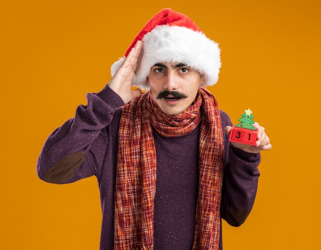 Wąsaty mężczyzna w świątecznej czapce świętego mikołaja z ciepłym szalikiem na szyi trzymający kostki zabawek z datą dwudziestu pięciu patrząc na kamerę zdezorientowany ręką nad głową stojącą na pomarańczowym tle