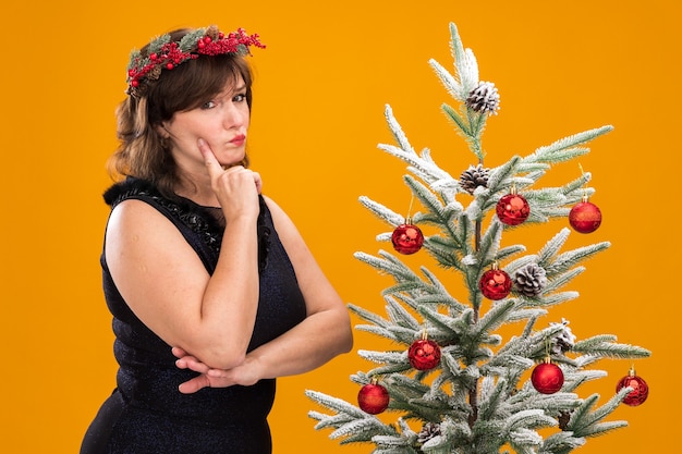 Wątpliwa kobieta w średnim wieku ubrana w świąteczny wieniec na głowę i świecącą girlandę na szyi stojąca w pobliżu udekorowanej choinki w widoku profilu dotykająca twarzy