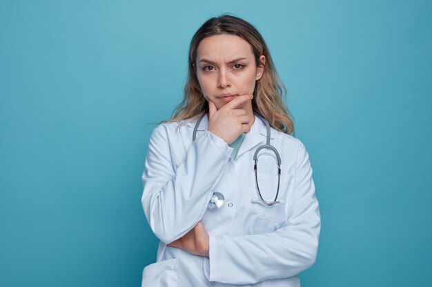 Wątpliwa młoda kobieta lekarz ubrana w szlafrok i stetoskop wokół szyi, trzymając rękę na brodzie