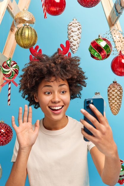 Wesoła Afroamerykanin macha ręką na ekranie smartfona, dzwoni do krewnych, zostaje w domu w okresie świąt Bożego Narodzenia, cieszy się przytulną atmosferą, która zdobi pokój przed zimą. Świąteczny nastrój