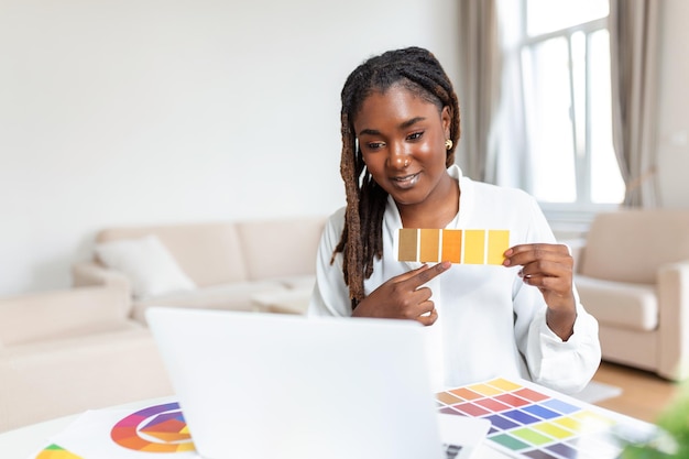 Wesoła afroamerykańska dama projektanta prowadząca wideokonferencję z klientami siedzącymi przy biurku przed komputerem, trzymająca palety kolorów, gestykulując i uśmiechając się do miejsca kopiowania