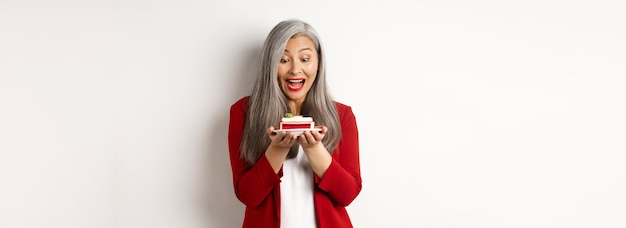 Bezpłatne zdjęcie wesoła azjatycka bizneswoman ciesząca się imprezą biurową, patrząca na pyszny kawałek ciasta i uśmiechająca się ul