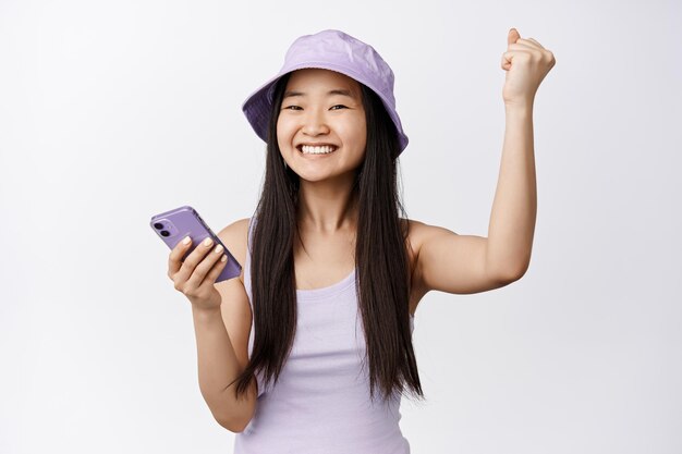 Wesoła azjatycka dziewczyna stojąca ze smartfonem i świętująca wygraną na telefonie komórkowym, uśmiechnięta i triumfująca stojąc na białym tle