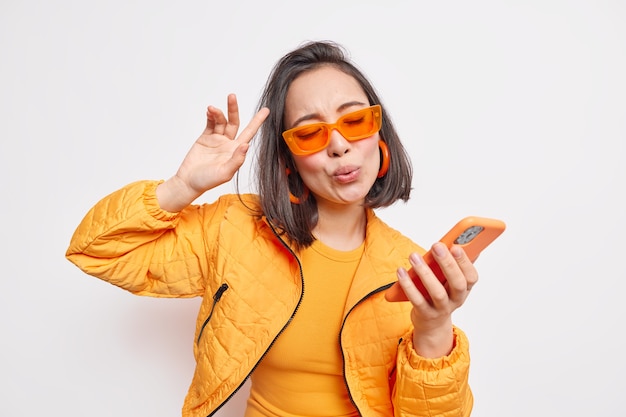 Wesoła brunetka Azjatka tańczy do ulubionej piosenki porusza się w rytm muzyki trzyma nowoczesny smartfon nosi modne pomarańczowe okulary stylowa kurtka wyraża szczęście i radość modeli w pomieszczeniach.