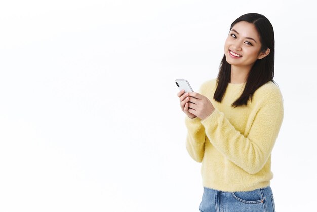Wesoła charyzmatyczna azjatycka kobieta uśmiecha się trzymając telefon komórkowy Dziewczyna wysyła wiadomość do przyjaciela za pośrednictwem smartfona edycja zdjęć za pomocą nowych filtrów aplikacji pobierz aplikację randkową lub nową grę aplikacji białe tło