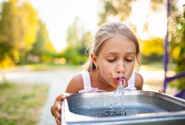 Wesoła cudowna dziewczyna pije chłodną świeżą wodę z małej fontanny w letnim ciepłym słonecznym parku na długo oczekiwane wakacje