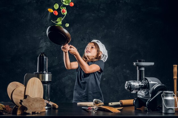 Wesoła dziewczyna rzuca warzywa na patelnię w ciemnym studiu fotograficznym.