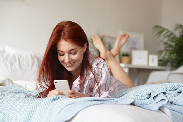 Wesoła figlarna młoda ruda kobieta w piżamie leżąca na łóżku w swoim pokoju z nogami w powietrzu, uśmiechająca się radośnie flirtując ze swoim chłopakiem, wysyłając mu SMS-a online za pomocą telefonu komórkowego