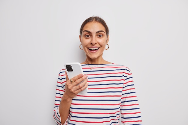 Wesoła kobieta o przyjemnym wyglądzie trzyma telefon komórkowy, cieszy się komunikacją online, nosi swobodny sweter w paski odizolowane na białej ścianie