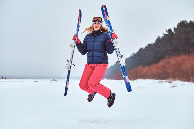 Wesoła kobieta ubrana w ocieplającą odzież sportową, trzymająca narty i skacząca na zaśnieżonej plaży