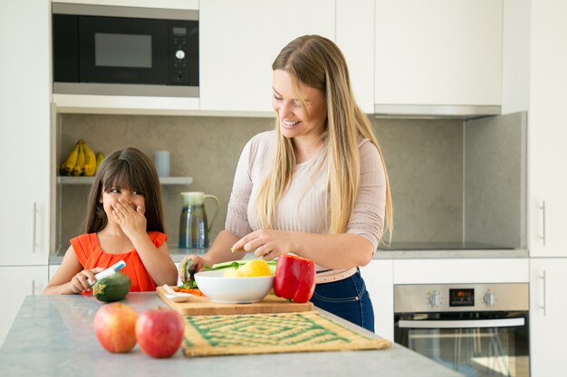 Wesoła mama i córka rozmawiają i śmieją się podczas gotowania warzyw na obiad. Dziewczyna i jej matka obierania i krojenia warzyw na sałatkę na blacie kuchennym. Koncepcja gotowania rodziny