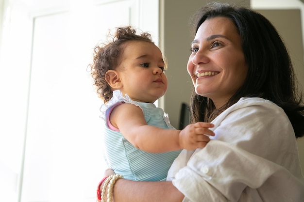 Wesoła mama trzyma w ramionach słodką córeczkę. Śliczne kręcone włosy dziewczynka patrząc na matkę. Koncepcja rodzicielstwa i dzieciństwa