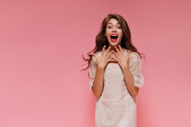 Bezpłatne zdjęcie wesoła młoda dziewczyna w białej sukni śmieje się i patrzy w kamerę ładna kobieta w stylowym stroju pozuje w zdziwionym nastroju na różowym tle