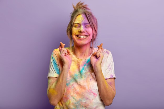 Wesoła młoda Europejka z zadowolonym wyrazem twarzy, skupiona na życzeniu, krzyżuje palce, brudna od kolorowego proszku Holi, ma nadzieję na fioletową ścianę, przychodzi na festiwal kolorów z przyjaciółmi.