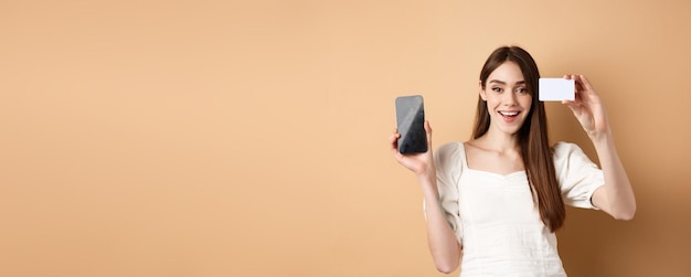 Wesoła młoda kobieta pokazująca plastikową kartę kredytową i pusty ekran telefonu komórkowego uśmiechająca się zadowolona z ok
