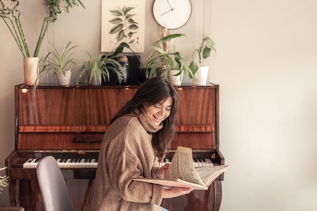 Wesoła młoda kobieta w przytulnym swetrze siedzi przy fortepianie i patrzy na nuty.