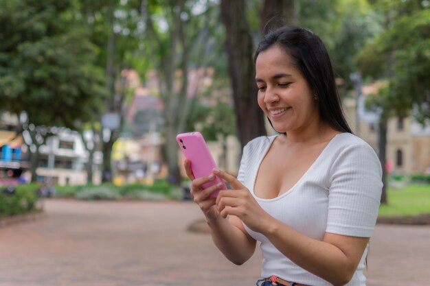 Wesoła młoda latynoska kobieta siedzi w parku i wysyła SMS-y przez telefon