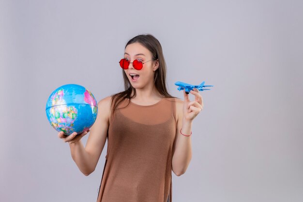 Wesoła młoda piękna kobieta nosi czerwone okulary przeciwsłoneczne, trzymając kulę ziemską i zabawkowy samolot, figlarny i szczęśliwy stojący