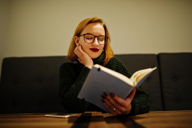 Bezpłatne zdjęcie wesoła młoda piękna rudowłosa kobieta w okularach nosi zielony ciepły wełniany sweter, siedząc w kawiarni i używając swojego notatnika
