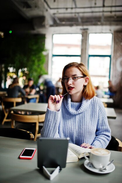 Wesoła młoda piękna rudowłosa kobieta w okularach za pomocą touchpada telefonu i notebooka, siedząc w swoim miejscu pracy w kawiarni z filiżanką kawy
