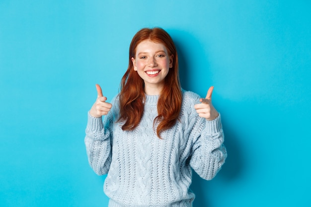 Wesoła nastolatka z rudymi włosami, wskazując palcami na aparat i uśmiechnięta, gratulująca lub chwaląca, stojąca na niebieskim tle