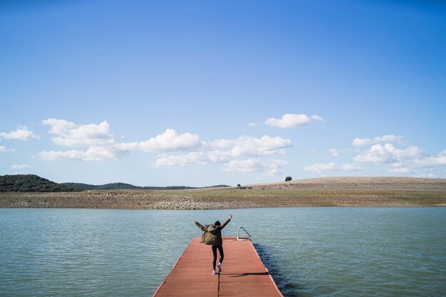 Wesoła osoba skacząca na przystani w pobliżu jeziora pod zachmurzonym niebem