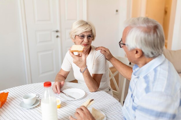 Wesoła para starszych jedząca śniadanie w domu Starszy mężczyzna przytula kobietę Dojrzała kobieta trzyma kawałek chleba Staruszek i kobieta siedzą przy stole rozmawiają