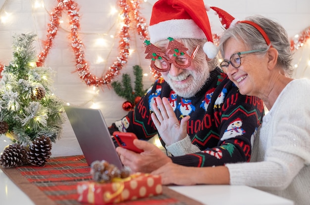 Wesoła para starszych w boże narodzenie słodsze i santa hat używa laptopa do rozmowy wideo z odległymi ludźmi. starsze pary świętują wydarzenie świąteczne. wesołych świąt i szczęśliwego nowego roku