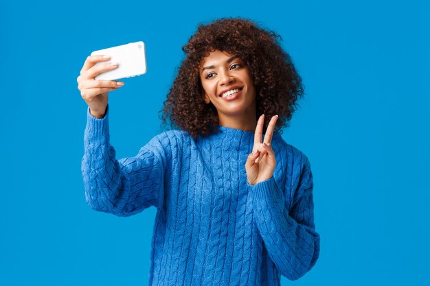 Wesoła, przyjazna i urocza afroamerykańska studentka, która sama robi zdjęcie, stosuje filtry w nowej aplikacji na smartfony, biorąc selfie przechylając głowę, uroczo uśmiechnięta, wykonująca gest pokoju, niebieska ściana.