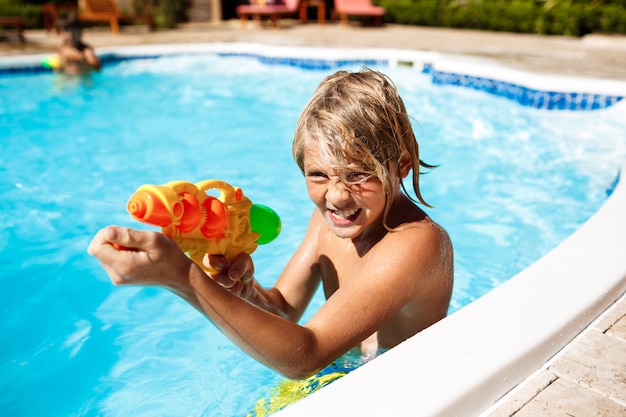 Wesołe dzieci bawiące się pistoletami wodnymi, radujące się, skaczące, pływające w basenie.