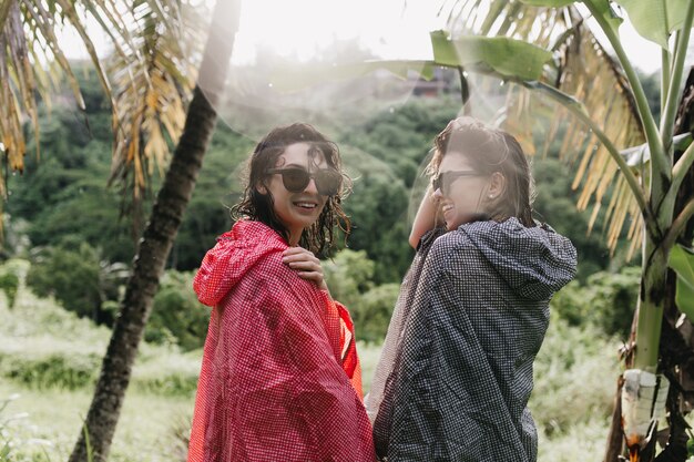 Wesołe kobiety chodzą po lesie w płaszczach przeciwdeszczowych. Odkryty strzał szczęśliwych koleżanek w okularach stojących na dżungli.