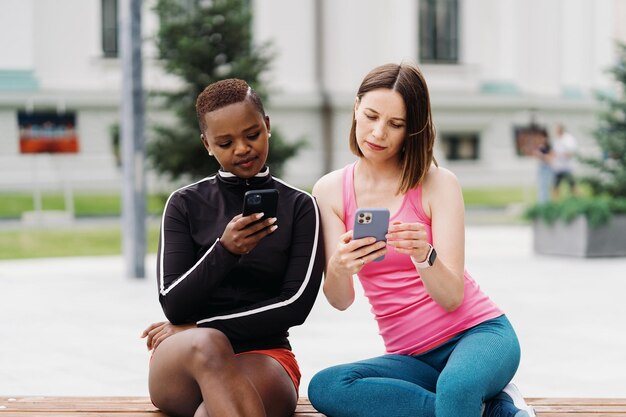 Wesołe uśmiechnięte koleżanki w strojach sportowych siedzą na ławce w mieście i rozmawiają podczas używania smartfona patrząc na ekran Wieloetniczne kobiety mają przerwę na trening fitness