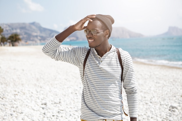 Wesoły afrykański młody człowiek stojący na plaży i patrząc na bok z rozkosznym uśmiechem zauważył zbliżającego się przyjaciela dotykającego głowy. Letnie wakacje i przygody. Ludzie, styl życia i podróże