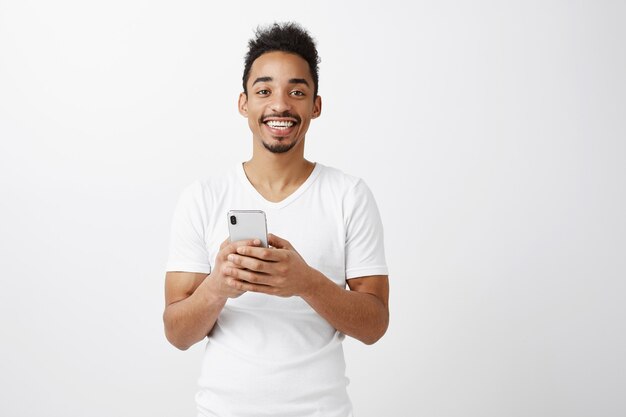 Wesoły atrakcyjny facet Afroamerykanin w białej koszulce trzymając telefon komórkowy, uśmiechając się