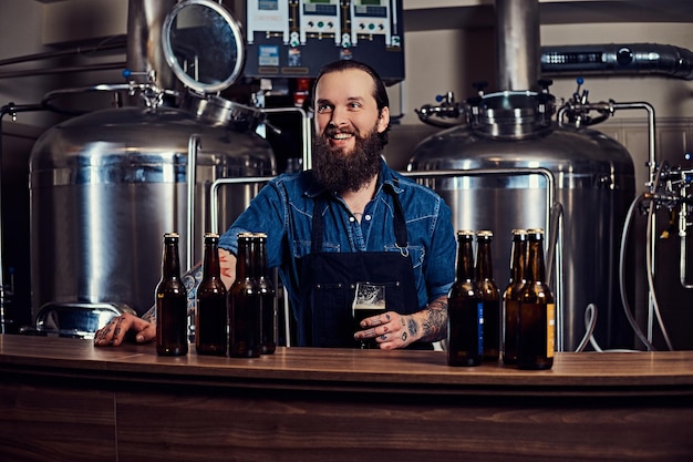 Wesoły, brodaty, wytatuowany mężczyzna hipster w dżinsowej koszuli i fartuchu, pracujący w fabryce browaru, stojący za ladą, trzyma kufel piwa do kontroli jakości.