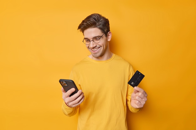 Wesoły facet o ciemnych włosach używa telefonu komórkowego i karty bankowej do robienia zakupów online, coś w sklepie internetowym wprowadza informacje, czy bezpieczna płatność na białym tle na żółtym tle