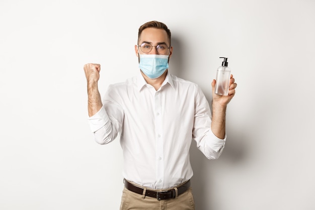 Wesoły kierownik w masce medycznej pokazujący środek dezynfekujący do rąk, stojący